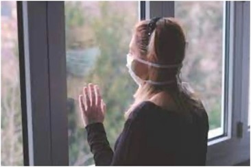 Woman wears a face mask while looking out of a window. She looks sad.